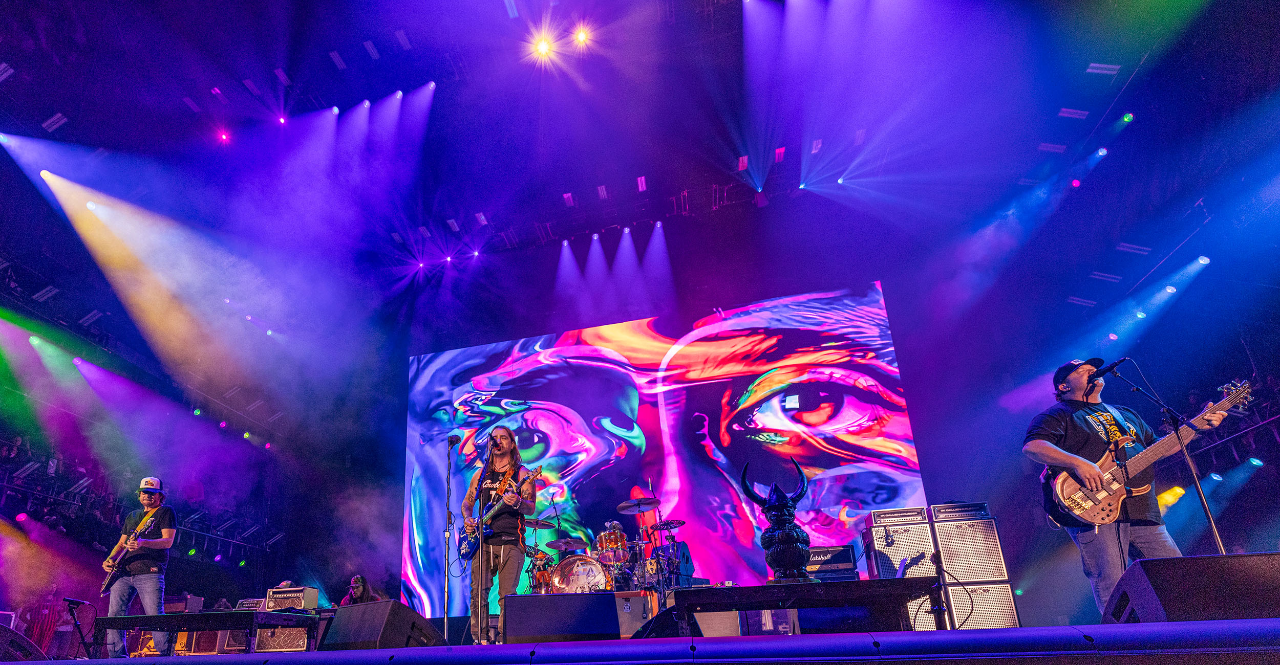 A vibrant concert stage with colorful lighting and a large, psychedelic backdrop featuring abstract human faces. The band members are playing their instruments, including guitars and drums, under the dynamic lights. The scene is energetic and visually captivating.