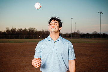 A male student on a baseball field tossing a baseball up to himself with a big smile on his face.