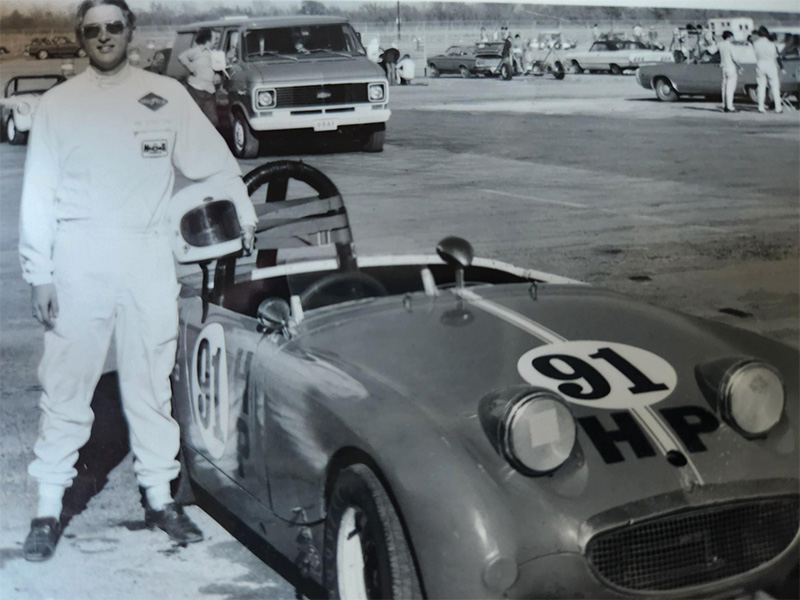 A black and white photo of a man in a racing suit next to a white racecar.