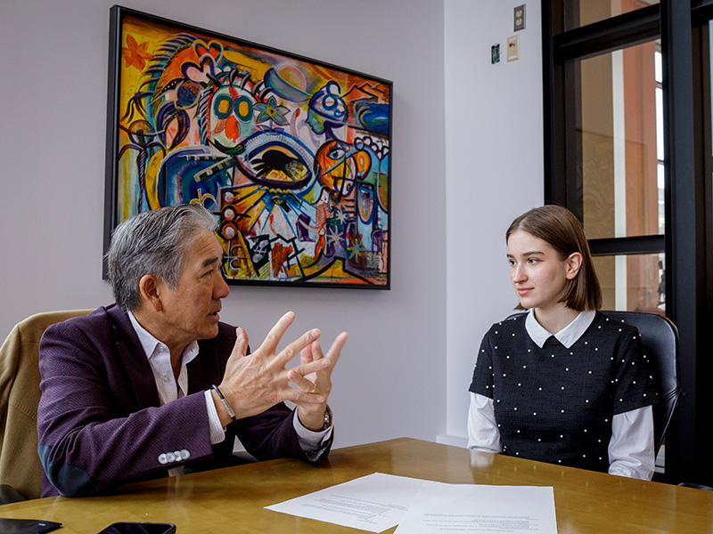 Two people seated at a wooden table in an office setting. One, with gray hair and a dark blazer, is gesturing with both hands while speaking. The other, with short hair and wearing a black sweater over a white collared shirt, is listening. Papers are spread on the table, and a colorful abstract painting hangs on the wall behind them.