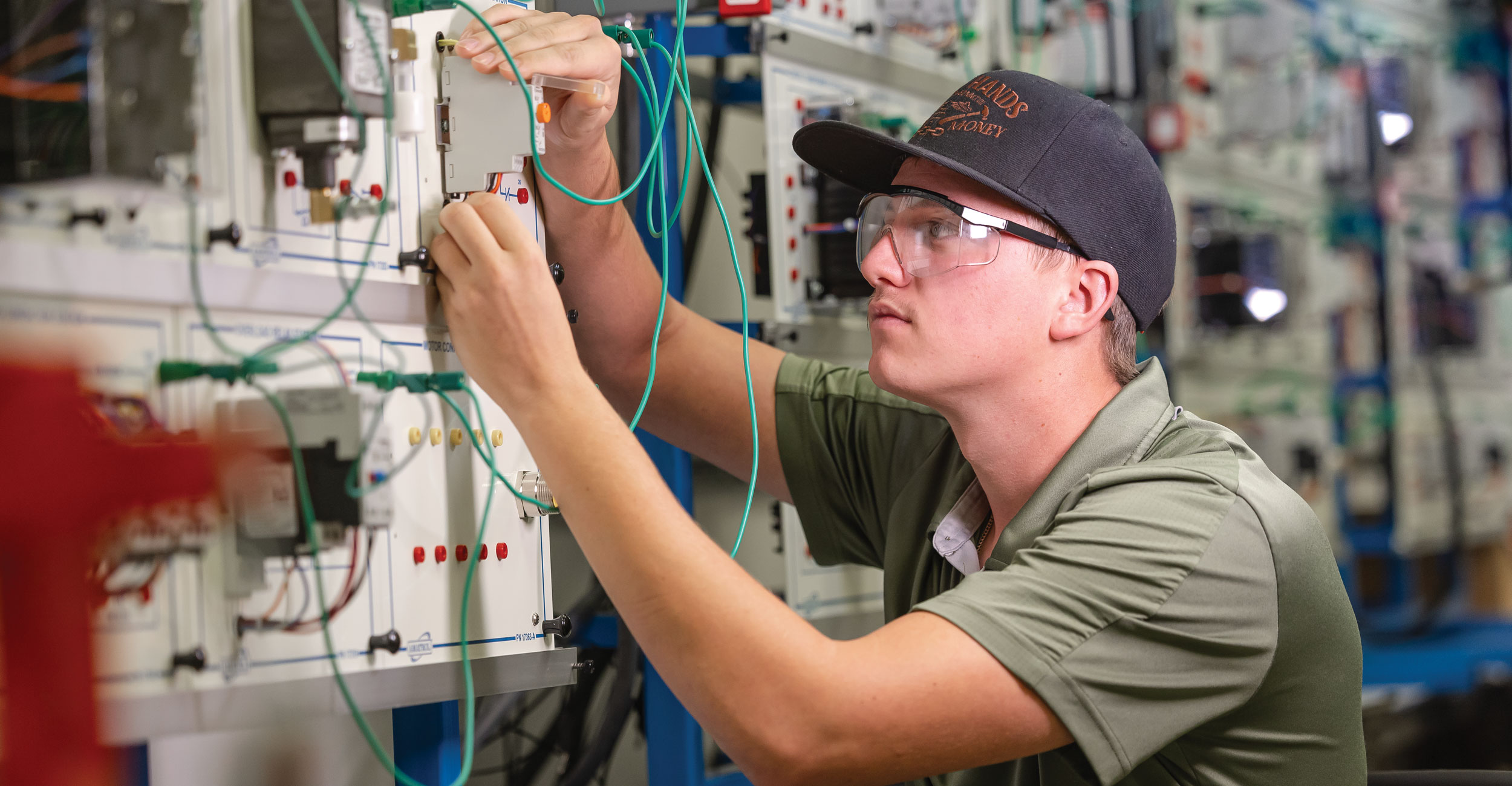 A man wearing a black hat and green shirt works with numerous wires on a electric box