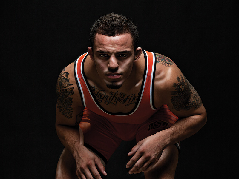 Jordan Oliver A muscular individual in an orange and black singlet crouches with both hands on their knees. Their face is blurred, and tattoos are visible on both shoulders and chest.