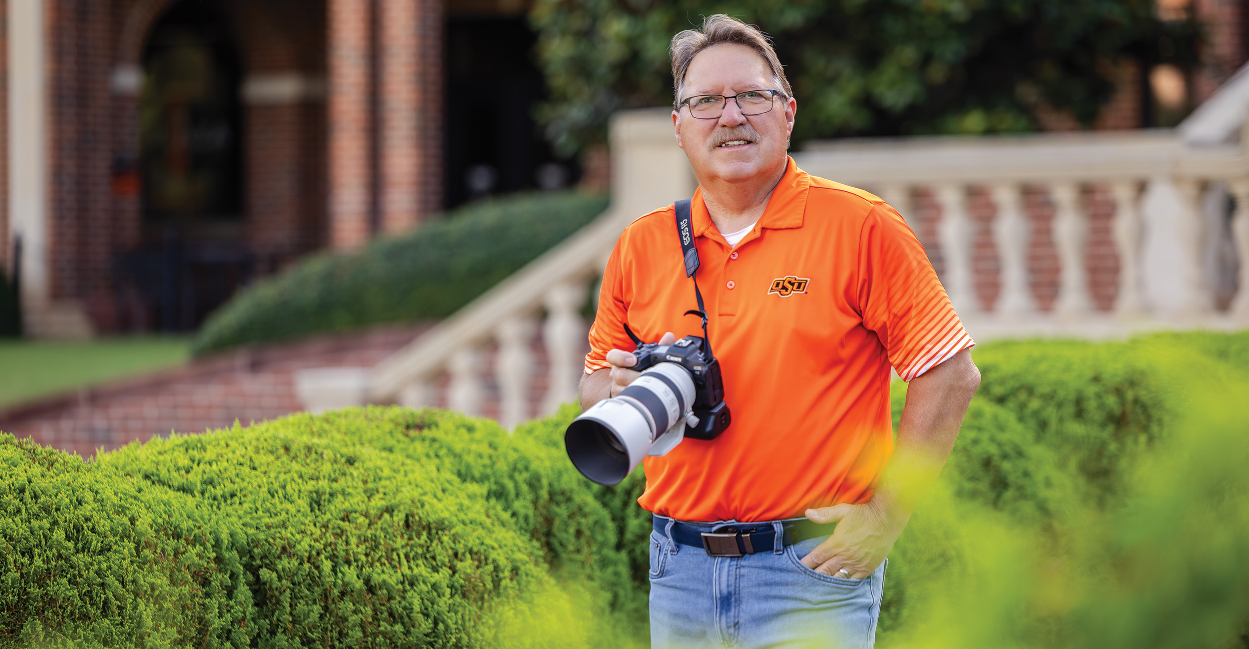 Gary Lawson A man in an orange shirt and blue jeans stands outdoors holding a professional camera with a large lens. The background includes green bushes, a brick building, and a stone railing.