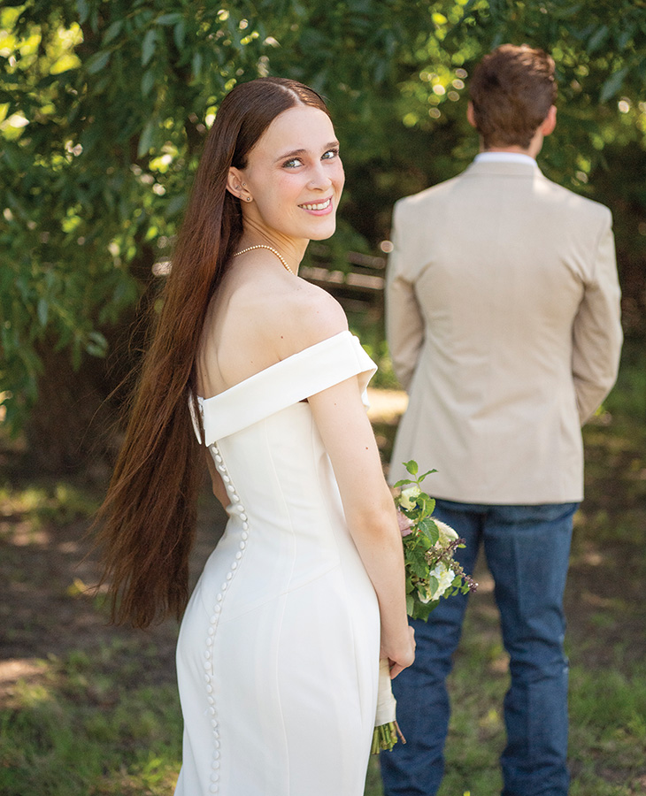 Jadrian Cook A woman in a white off-shoulder wedding dress with long brown hair stands outdoors holding a small bouquet of flowers.