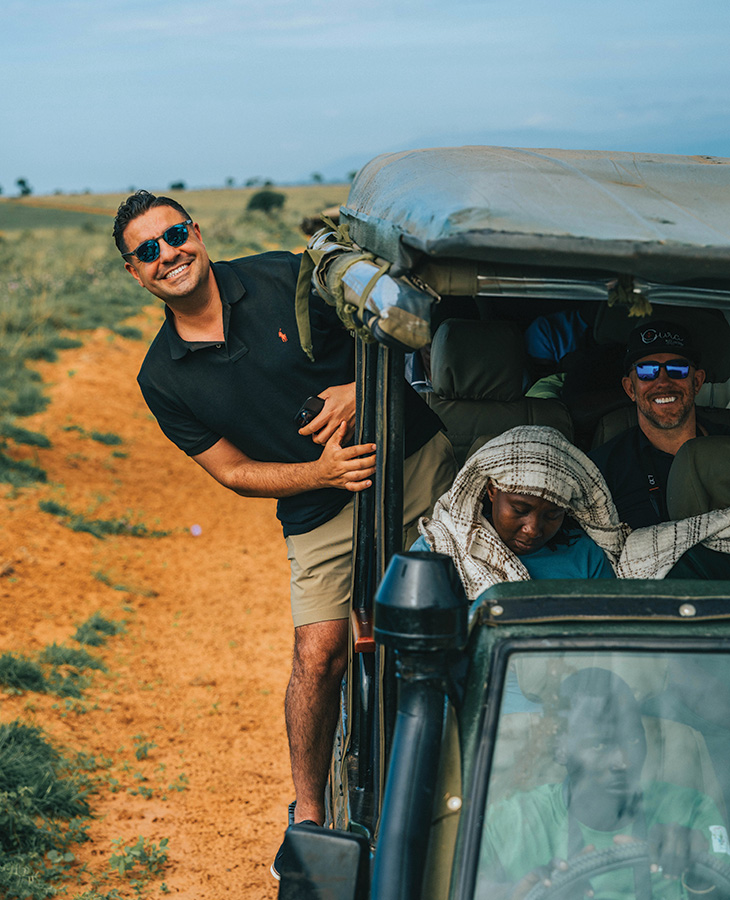 TJ Trad A man in a black shirt stands outside of a vehicle as it is driving on a dirt road.