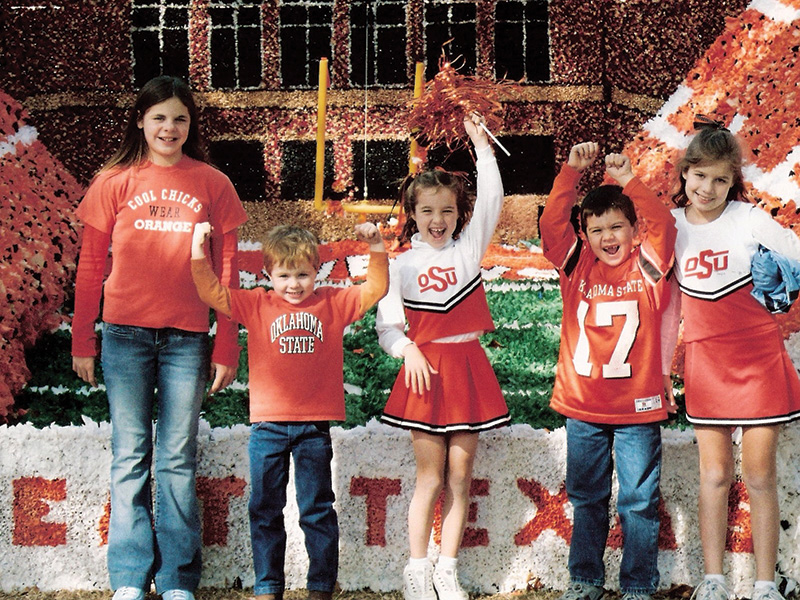 Nickles Family Five children dressed in Oklahoma State University (OSU) cheerleading and sports attire stand in front of a float decorated with an image of a building and the words 'GO POKES' spelled out in flowers.