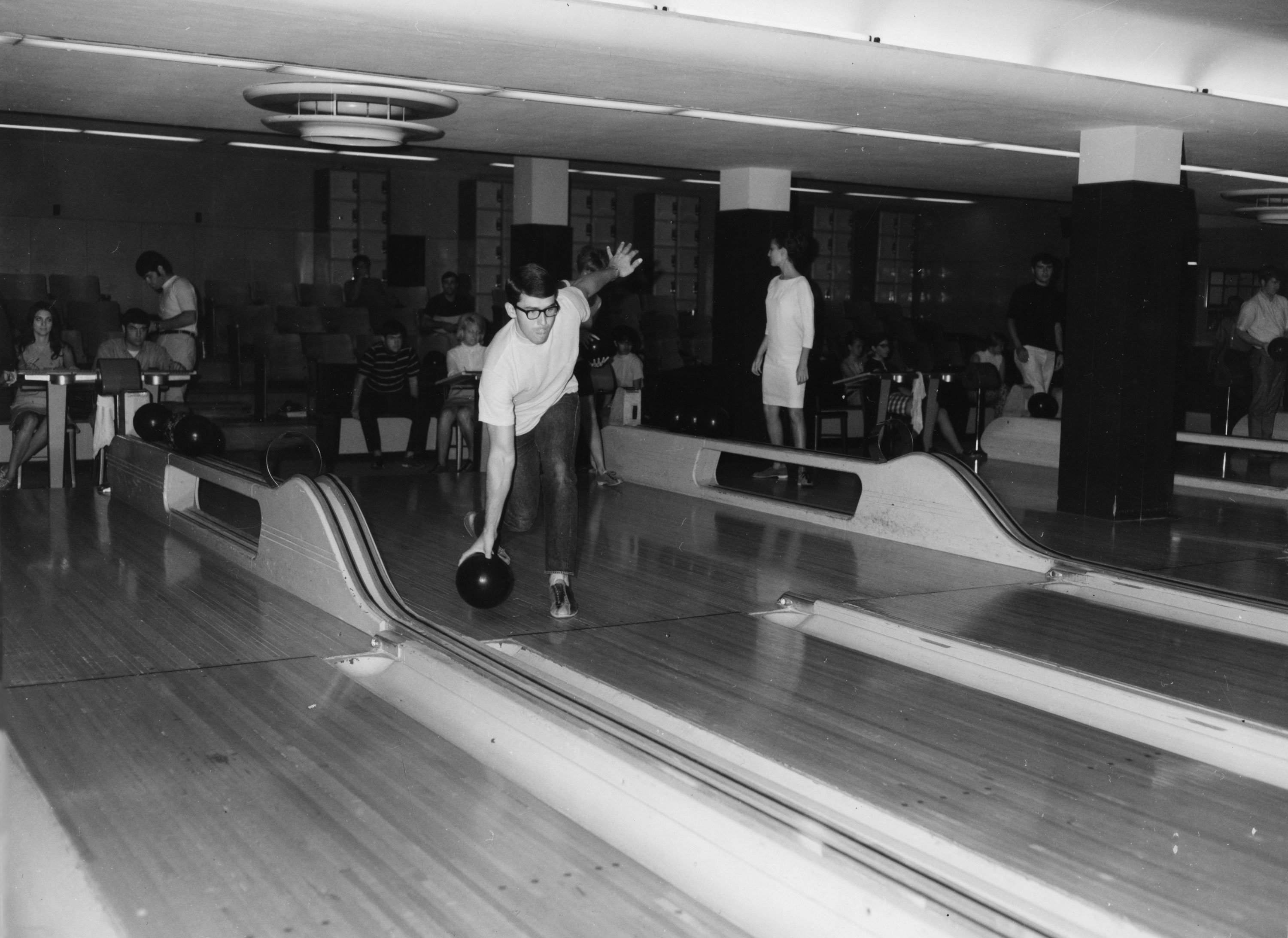 Bowling A black and white photograph of a bowling alley with multiple lanes. In the foreground, a man wearing glasses and a white shirt is in mid-action, preparing to release a bowling ball.