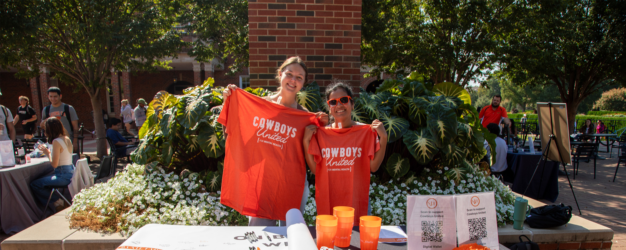 Cowboys United Outdoor event with two people holding bright orange T-shirts that read ‘Cowboys United’ in front of a table displaying orange cups, flyers, and QR codes, with greenery and a brick wall in the background.