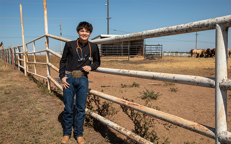 Alexander Nunez Person wearing a dark long-sleeve shirt with a large decorative belt buckle, standing outdoors beneath a wooden archway with a metal OSU logo, in front of a light-colored building and green shrubs under a clear blue sky.