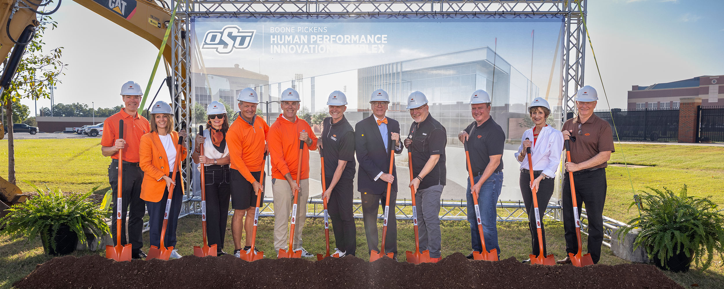 BPHPIC Groundbreaking A group of people wearing hard hats stand in a line holding orange ceremonial shovels at a groundbreaking event. They are positioned in front of a banner displaying the OSU logo and the words ‘Boone Pickens Human Performance Innovation Complex,’ with a rendering of the future facility shown behind them. The group stands on a lawn beside construction equipment and decorative plants under a clear sky.