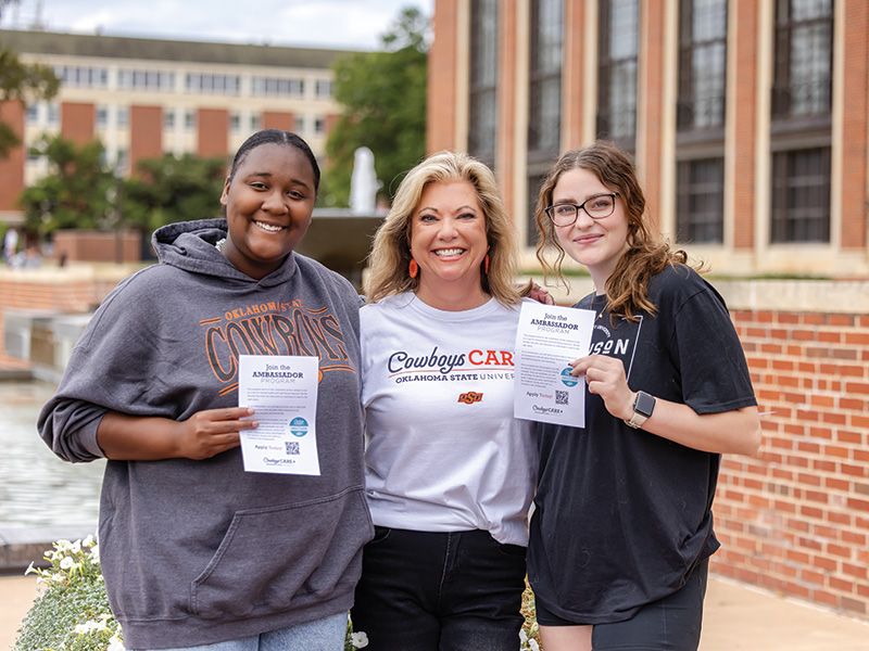 Three people standing outdoors near a brick building and fountain, wearing casual clothing and holding ambassador program flyers; one shirt reads ‘Cowboys Care Oklahoma State University.’