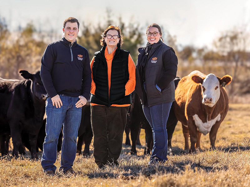 A man and two women stand in a field in front of some cattle.