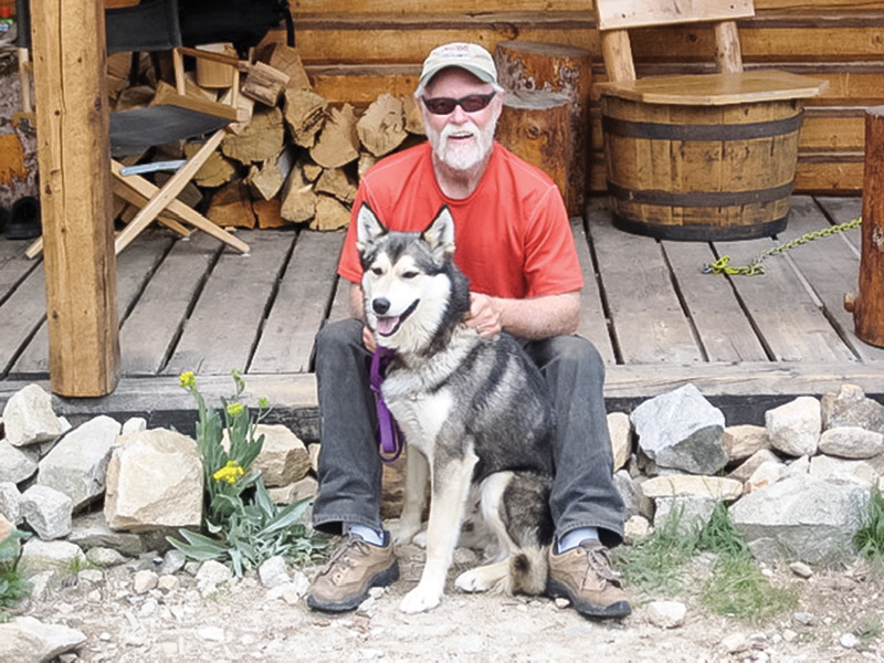 A person wearing a red T-shirt, dark pants, and hiking shoes sits on a wooden porch in front of a log cabin, with a black-and-white husky sitting between their legs. Stacked firewood and rustic furniture are visible in the background.